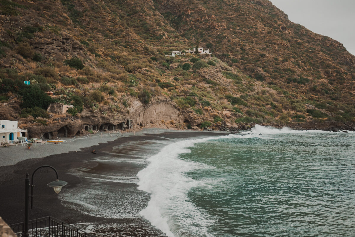 Schwarzer Sandstrand mit welligem Meer und Bergpanorama.