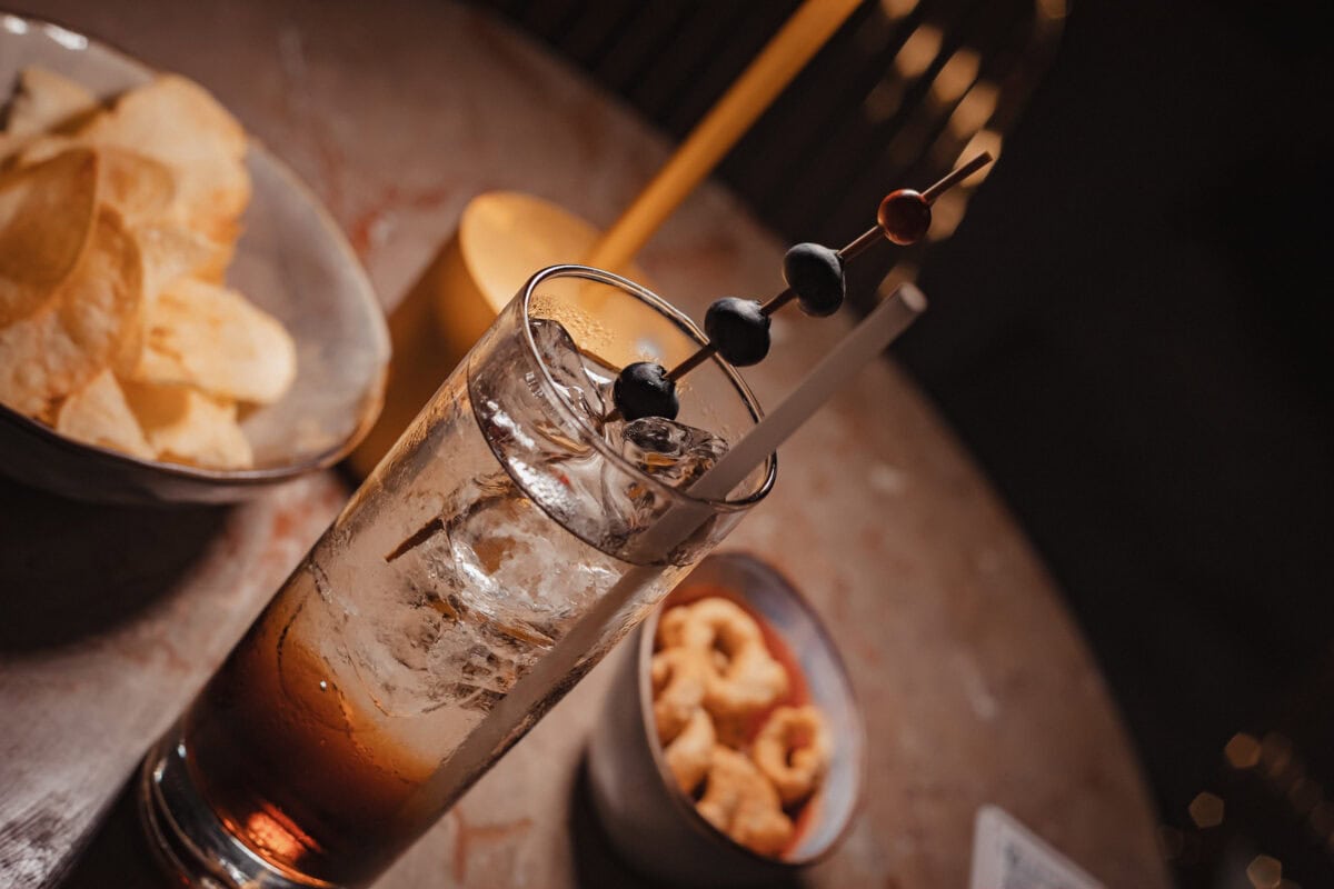Cocktailglas mit Eis und Oliven, Snacks im Hintergrund.