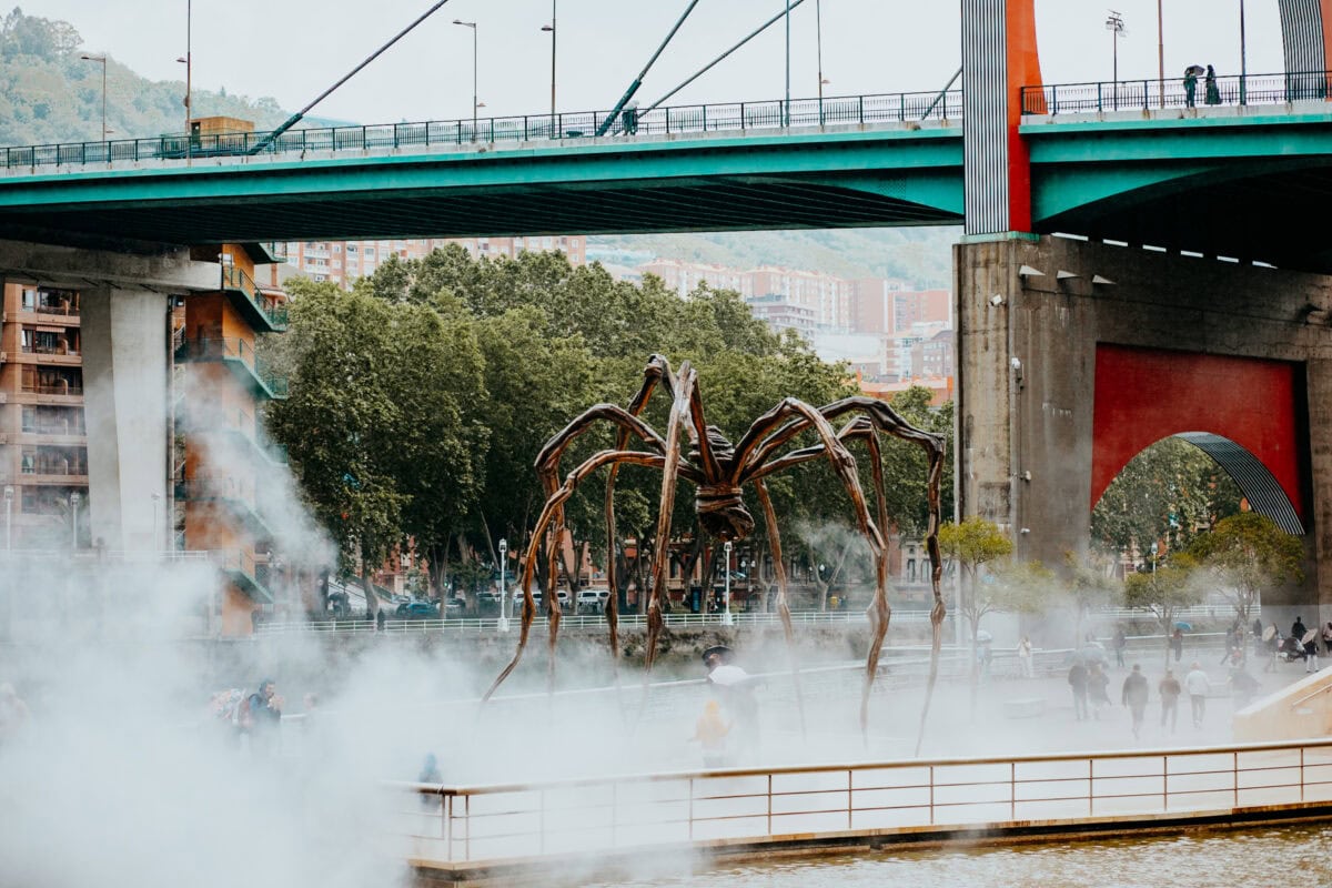 Metallische Spinnen-Skulptur im Nebel vor Brücke.