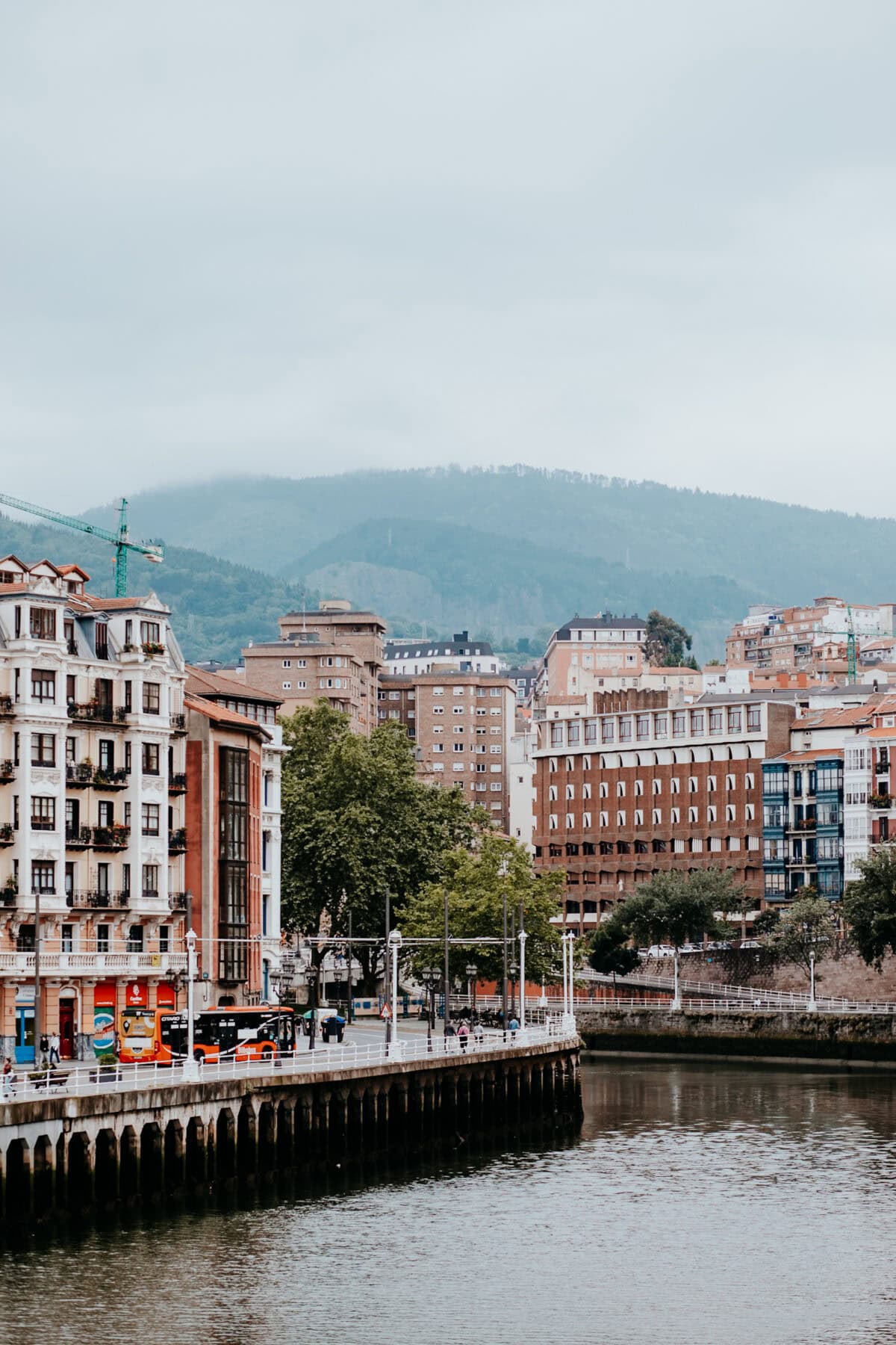 Stadtansicht mit Fluss und bewaldeten Hügeln im Hintergrund.