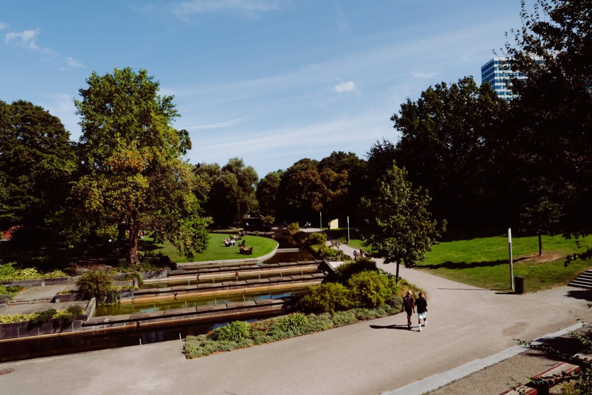 Sonniger Stadtpark mit Wasserbecken, Bäumen und Spaziergängern