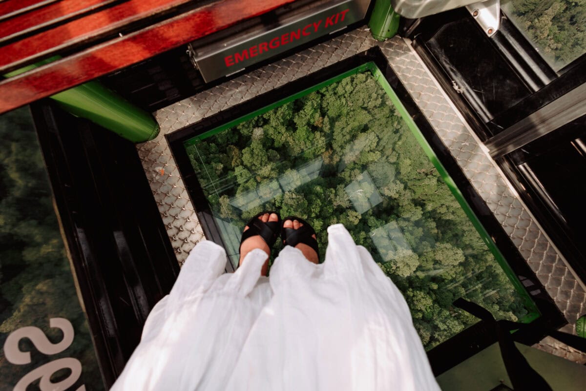 Füße auf Glasboden einer Seilbahn über grünem Wald